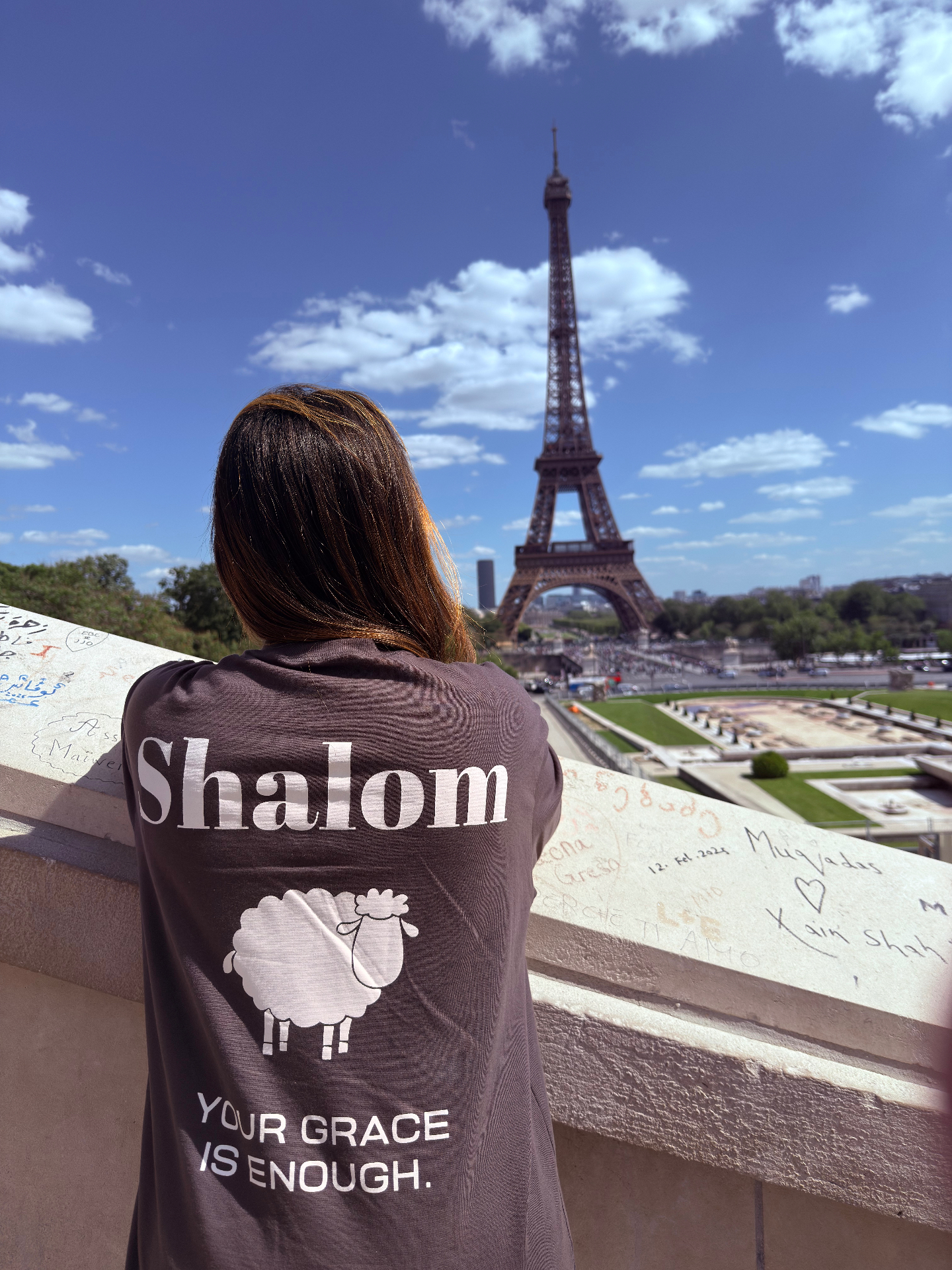 Mulher usando camiseta marrom SHALOM Your Grace is Enough em frente à Torre Eiffel em Paris